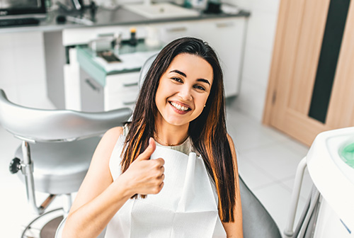 A woman is sitting in a dental chair, smiling and giving a thumbs-up gesture.